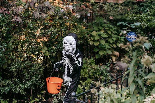 Frightening child in skeleton costume walking along garden with bucket