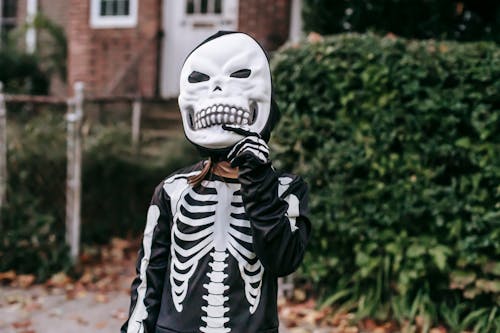 Crop faceless kid dressed in skeleton costume taking off scary mask while standing near house with fallen leaves and foliage on Halloween day