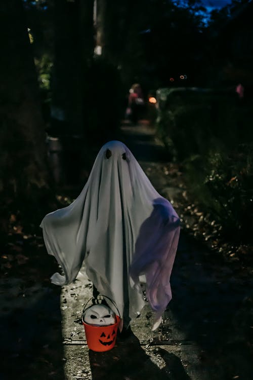 Anonymous kid dressed like frightening ghost standing alone on dark street between greenery with white mask in jack o lantern bucket during Halloween celebration