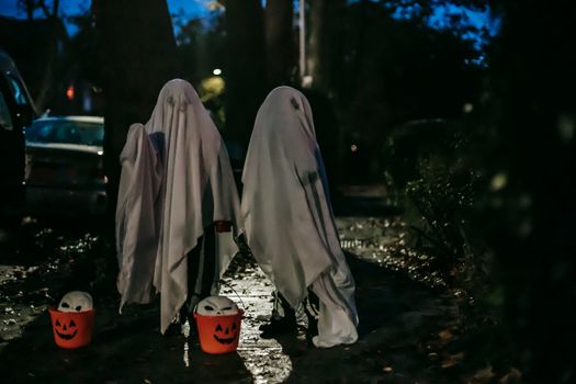一个无法辨认的孩子站在黑暗的街道上，用水桶捣蛋，身上盖着白色毯子，像鬼魂一样出现在万圣节庆祝活动中