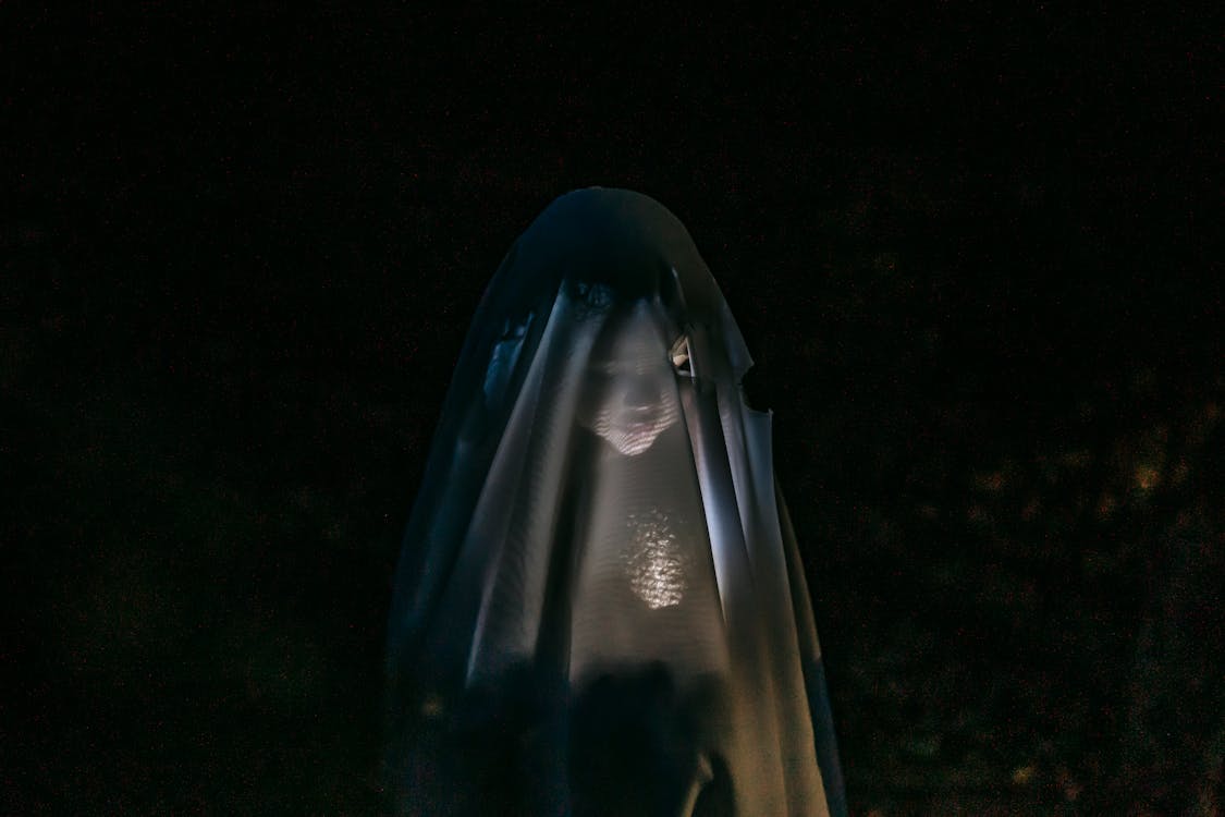 Free Mysterious little girl standing in darkness covered with white blanket as ghost and shining flashlight on face Stock Photo