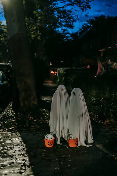 Unrecognizable kids in Halloween ghost costumes standing in shadow of illuminated tree