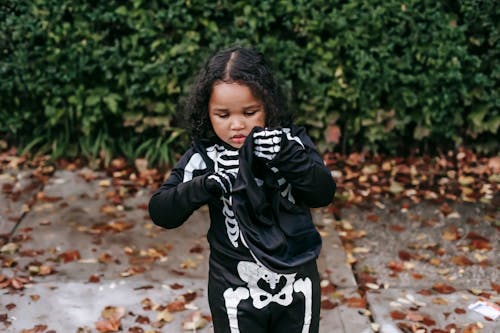Concentrated dark haired curly African American female child in skeleton costume putting on mask in Halloween in autumn park on pathway