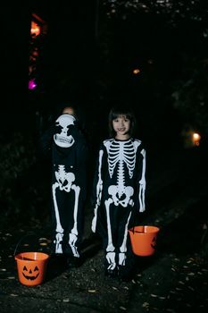 两个穿着骷髅服装的孩子，晚上拿着南瓜桶庆祝万圣节