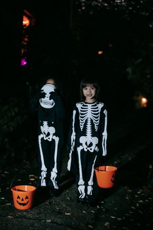 Free Full body creepy kids in skeleton costumes with orange pumpkin buckets with candies standing on street at night during Halloween celebration Stock Photo