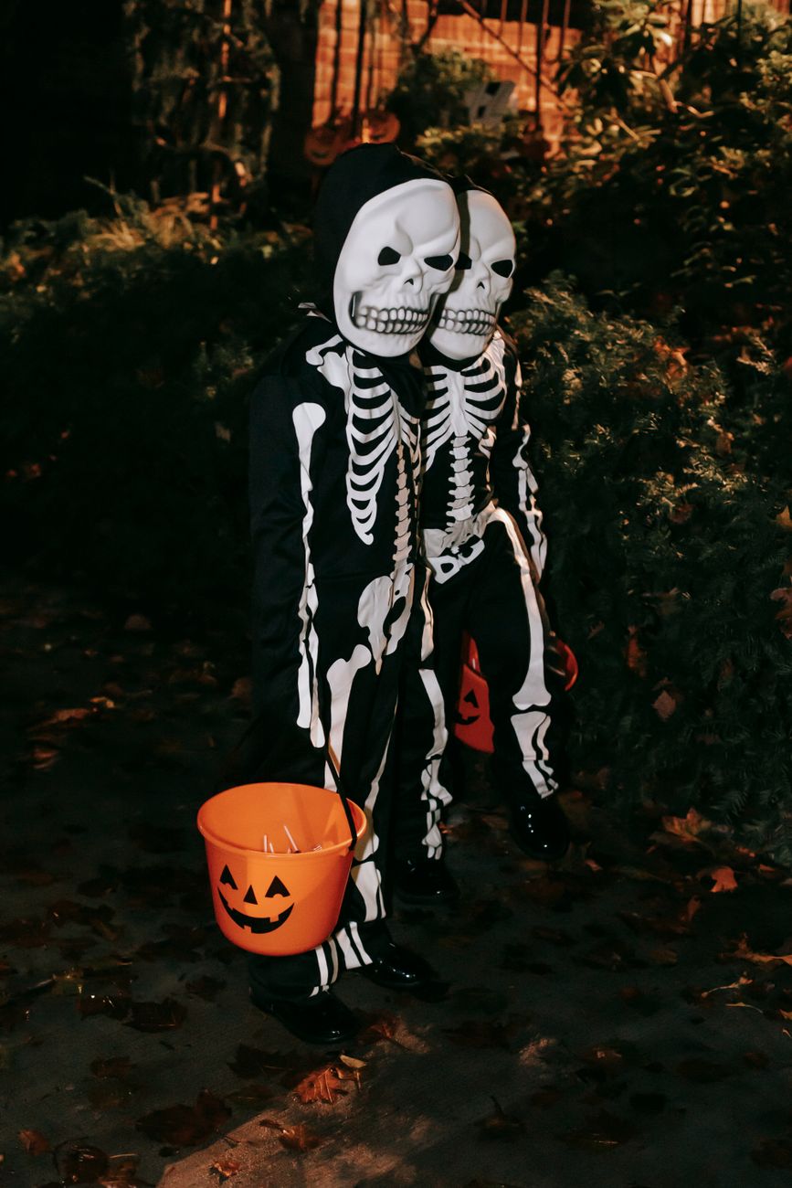 Two people in skeleton costumes holding orange pumpkin buckets in a park on Halloween night.