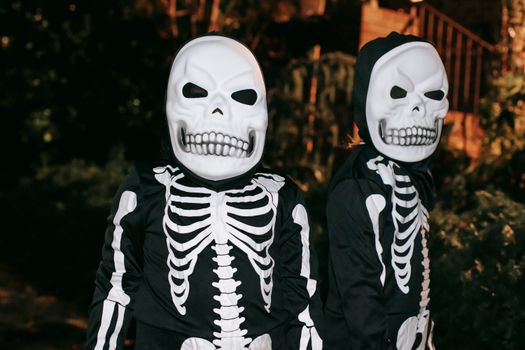 Creepy unrecognizable persons in skeleton costumes and skull masks standing together on dark street at night time during Halloween celebration