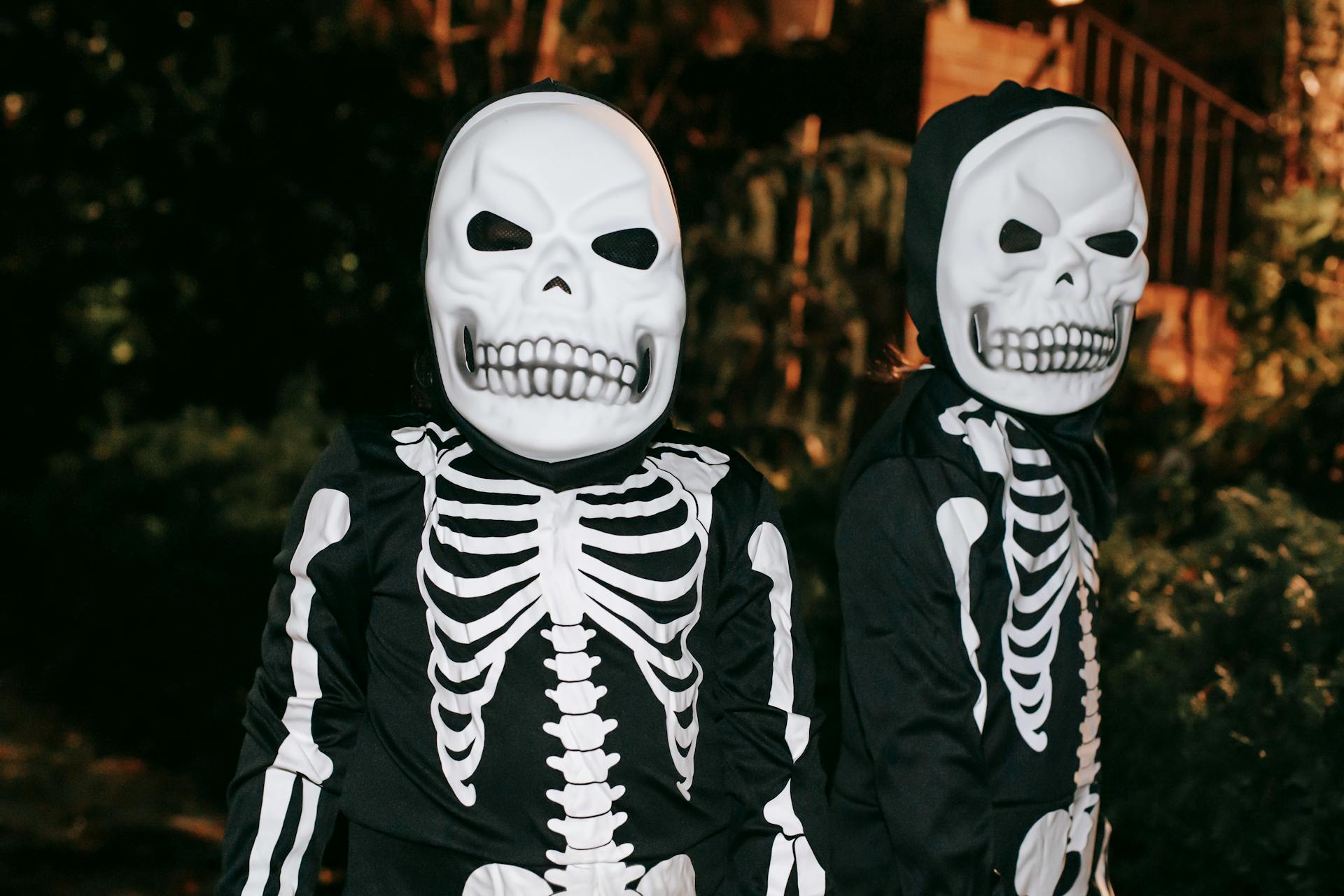 Creepy unrecognizable persons in skeleton costumes and skull masks standing together on dark street at night time during Halloween celebration