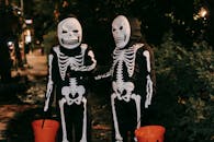Funny unrecognizable kids in skeleton costumes and masks with orange buckets in hands communicating to each other on street at Halloween night