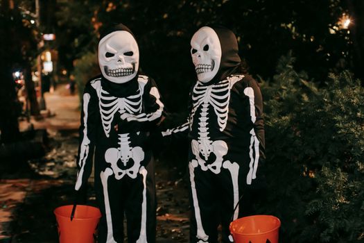 Funny unrecognizable kids in skeleton costumes and masks with orange buckets in hands communicating to each other on street at Halloween night