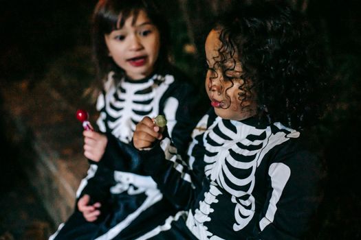 Crop interested girl in skeleton costume interacting with best African American friend with sucker during Halloween holiday in twilight