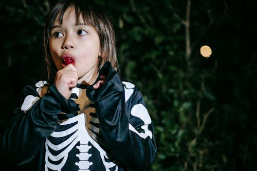 Crop contemplative girl in skeleton costume eating delicious lolly while looking away during festive occasion in park in twilight