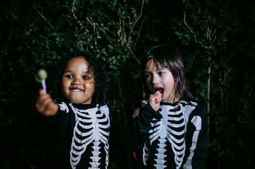 Content best multiracial friends in skeleton costumes eating tasty lollies during festive event at dusk outdoors