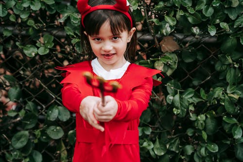 Crop adorable ethnic girl wearing red devil costume and pointing trident at camera while standing in park