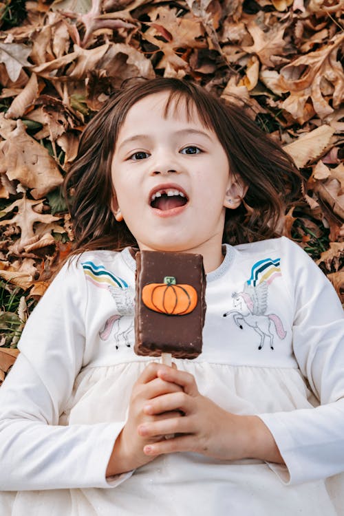 From above happy little girl enjoying chocolate ice cream decorated with pumpkin while lying on fallen autumn leaves and looking at camera merrily