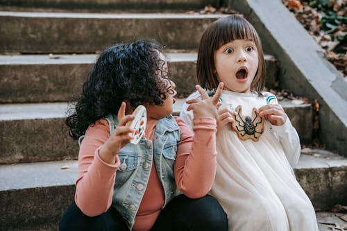 Amazed diverse girls with Halloween gingerbread cookies sitting on staircase