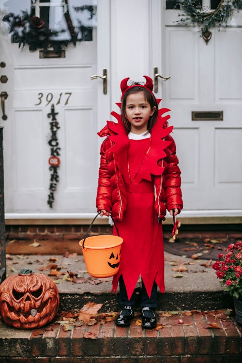Full body positive little girl wearing red devil costume with Trick or Treat bucket standing on doorsteps with Halloween decorations and looking at camera