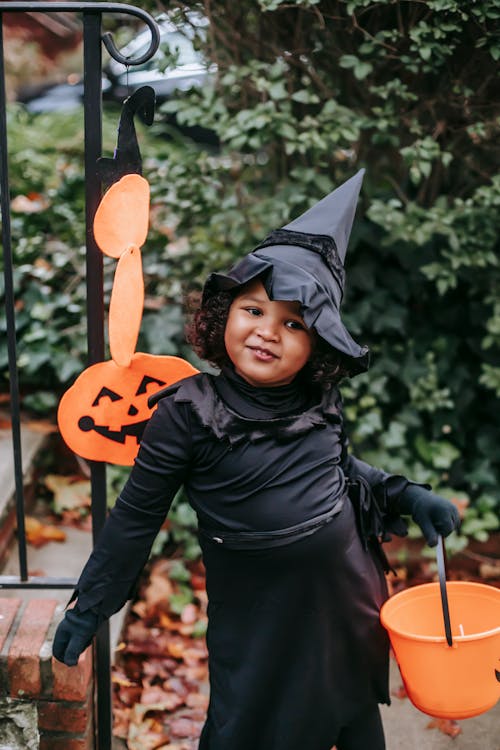 Happy little ethnic girl wearing black witch costume standing with Trick or Treat bucket in autumn garden and looking away contentedly