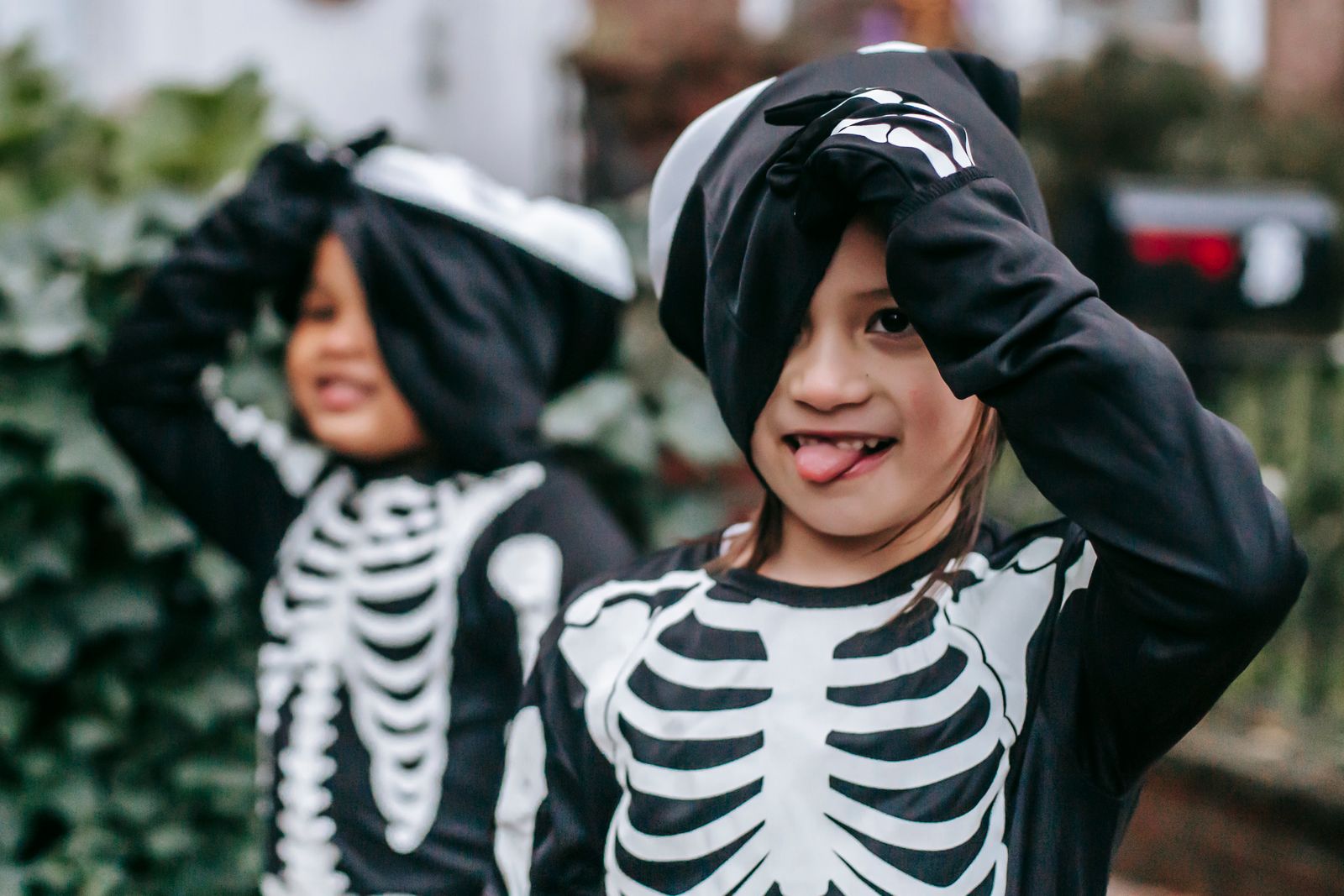 Two smiling children in matching skeleton Halloween costumes playing on a leafy residential street in autumn