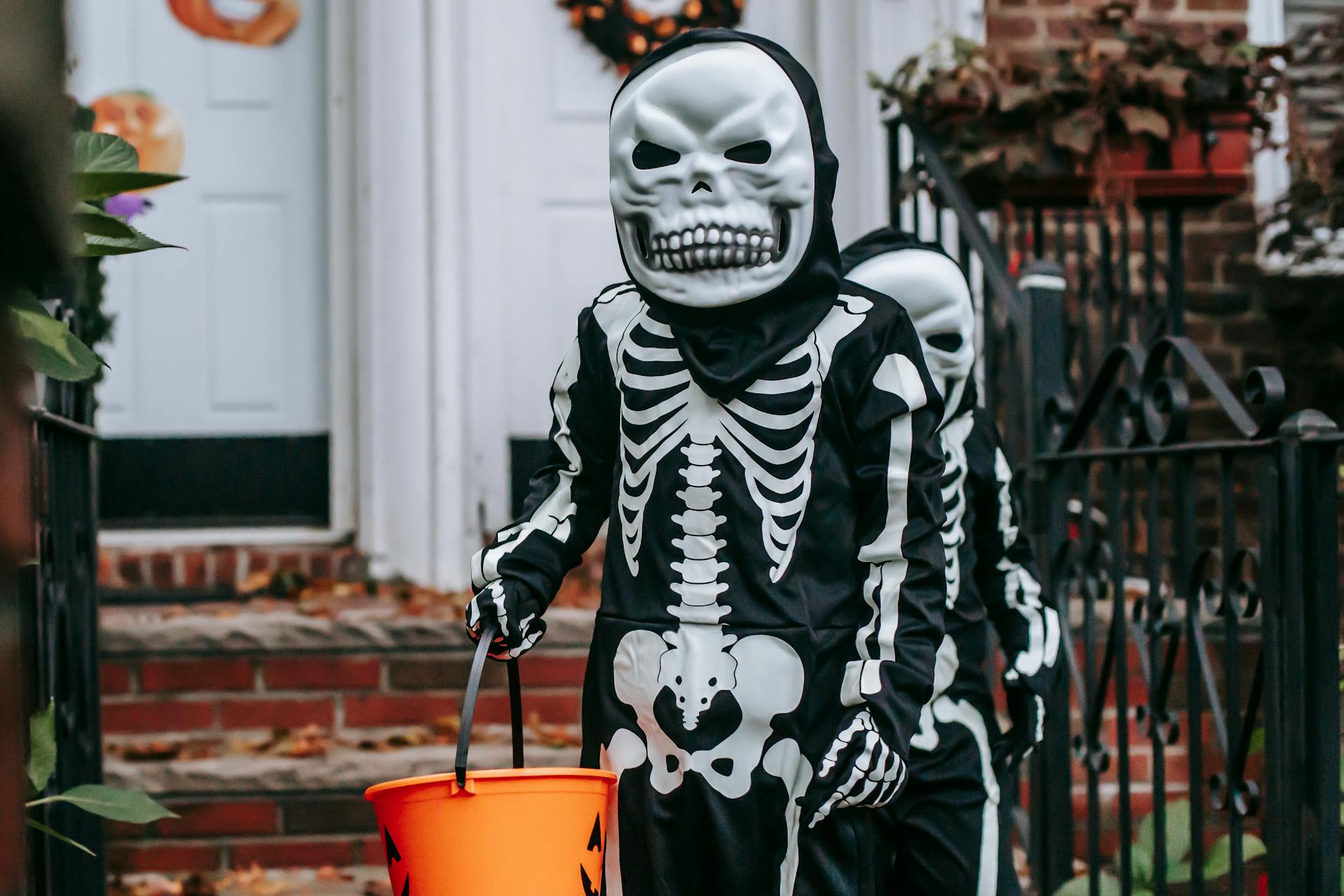 Anonymous kids with buckets wearing skeleton costumes and masks standing on terrace near entrance of house during trick or treat game