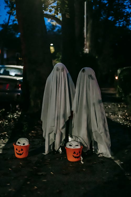 Free Full body of anonymous kids wearing white ghost costumes standing near tree trunk and orange buckets while playing trick or treat on street in evening Stock Photo