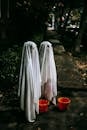 Full body side view of anonymous kids wearing white ghost costumes standing on sidewalk with orange buckets while playing trick or treat in evening