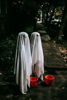 一个穿着白色幽灵服装的匿名孩子，夜晚站在人行道上玩“不给糖就捣蛋”游戏，手里拿着一个橙色水桶