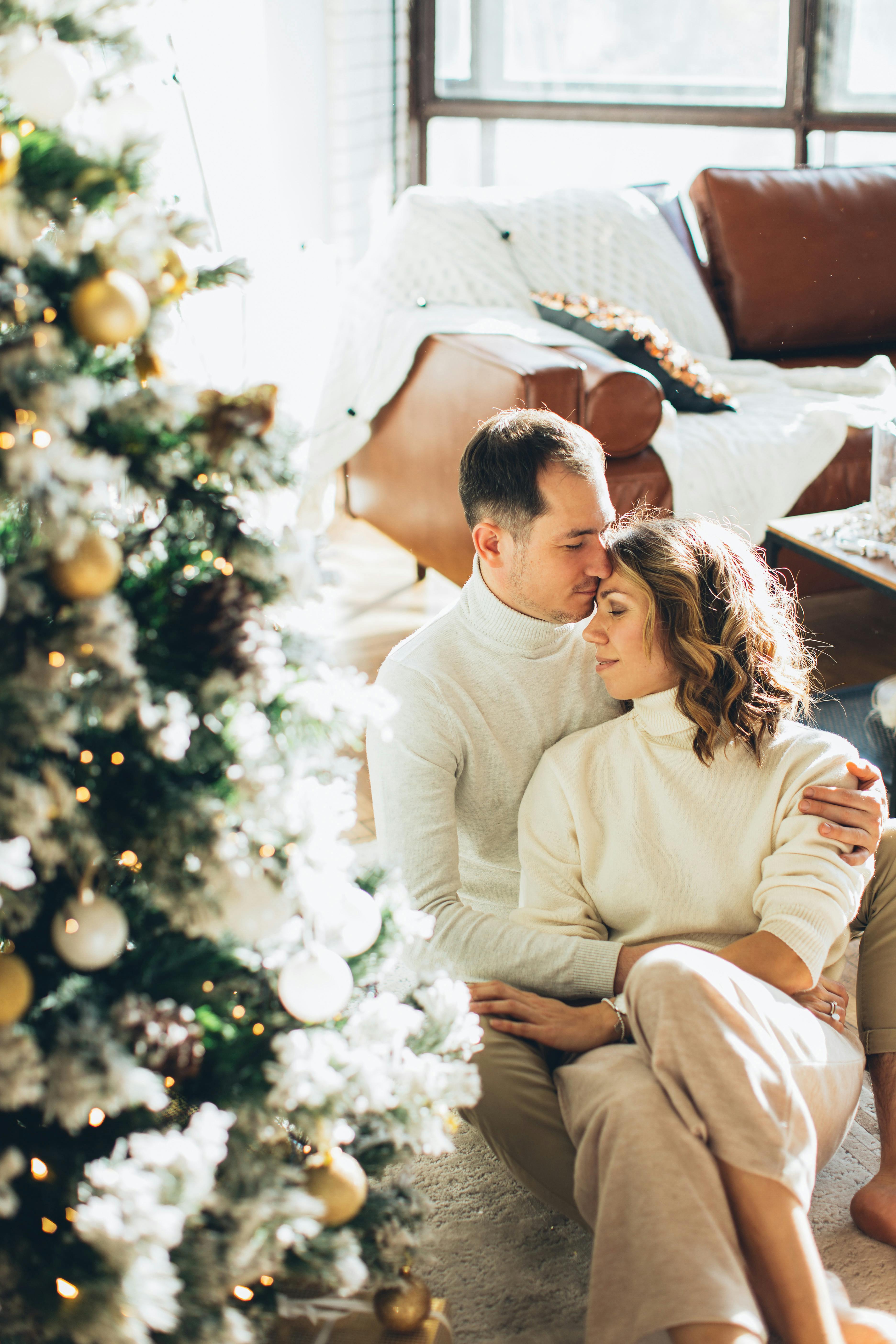 Couple in white sweaters hugging by a Christmas tree in a cozy room setting.