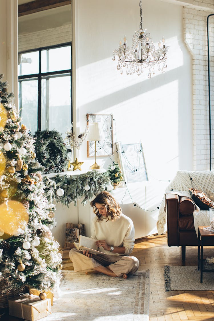 Woman In White Long Sleeve Shirt Sitting Near A Christmas Tree Reading A Book