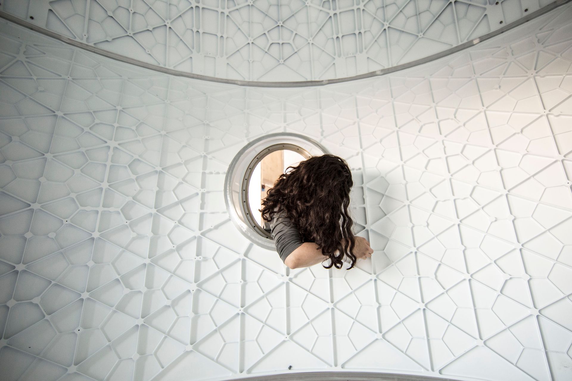 A woman peers through a circular opening in a modern geometric structure, adding a sense of curiosity.