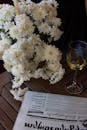 White Flowers Beside a Glass of Wine and Newspaper on Wooden Table