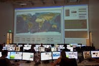 在飞行控制室里，一名无法识别的员工背对镜头，正操作计算机分析数据，墙上挂着一幅大型交互式地图