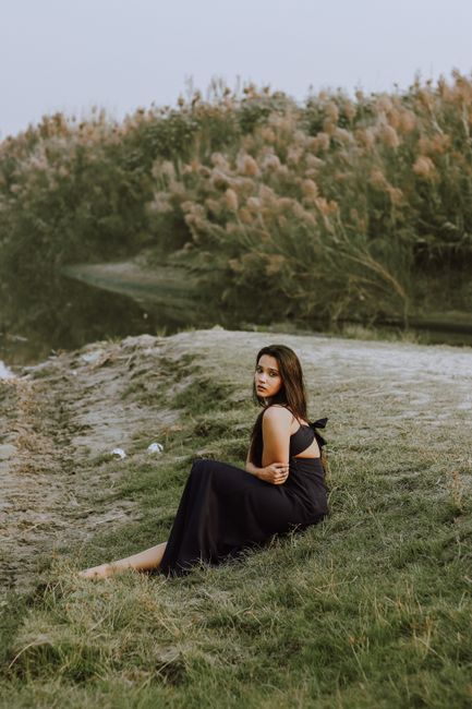 Full body of charming ethnic female in elegant outfit looking at camera while sitting with bent leg in nature near calm water