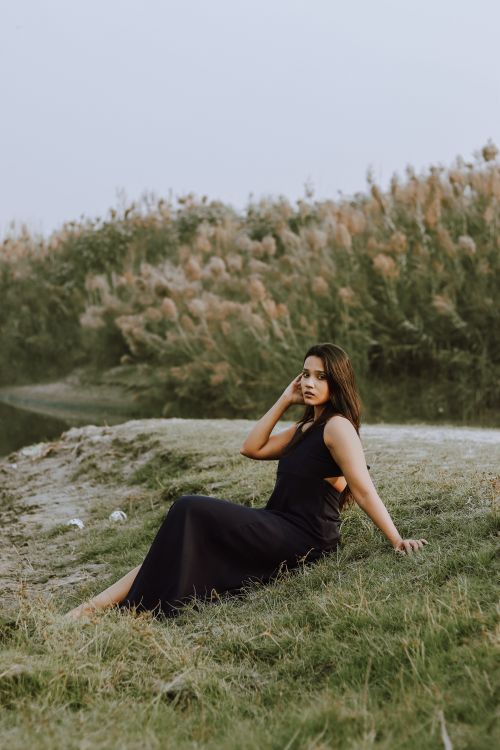 Free Full body of stylish ethnic female in black dress looking at camera and touching hair while sitting on green coast near lake Stock Photo