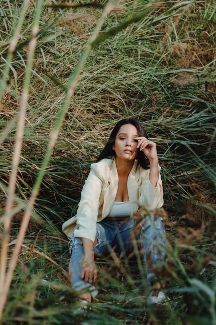 Stylish ethic female in trendy blazer leaning forward and looking at camera while sitting with bent arm on grass in rural area