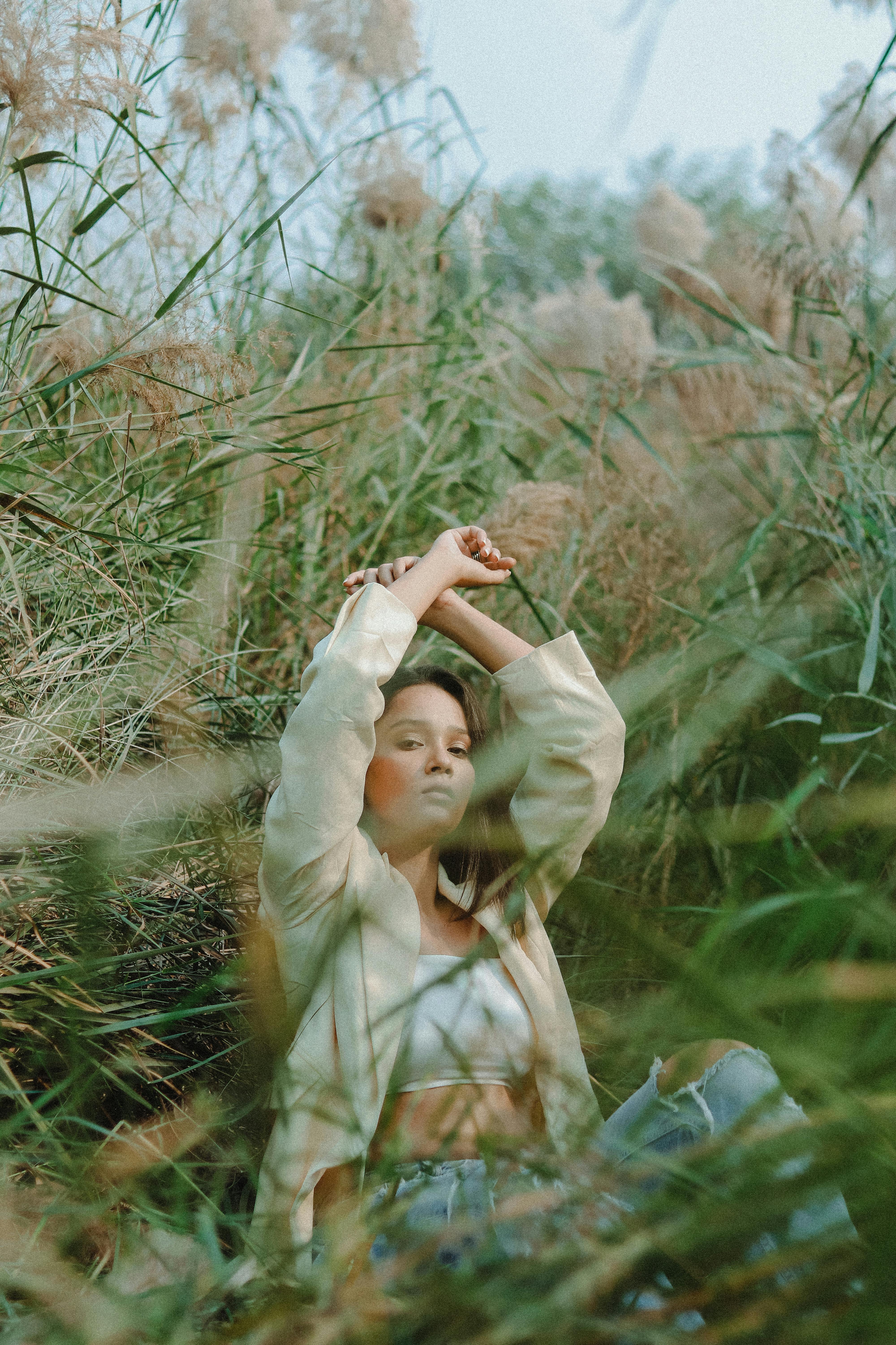 Stylish ethnic female with raised arms looking at camera while sitting on grassy ground near lush shrubs in summer nature