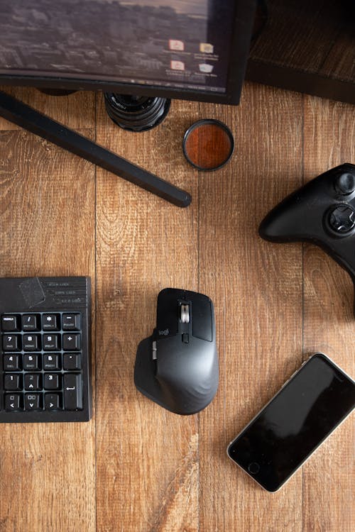 From above of black smartphone with keyboard and gamepad on wooden desktop with monitor in soft daylight at home