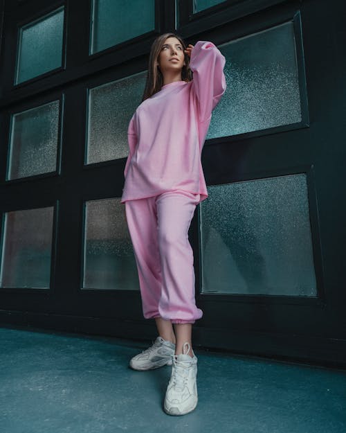 Low angle of female in trendy pink loose outfit standing at wall with frames and looking away