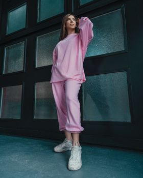 一位穿着时尚粉色宽松服装的女性，正低角度站在带镜框的墙边，目光移向别处