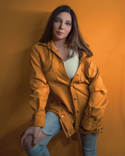 Stylish young female with fair hair in yellow jacket and ripped jeans looking at camera in soft daylight