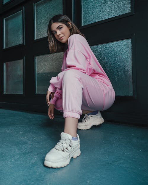 Full body of cheerful ethnic stylish woman in pink outfit and white sneakers hunkering down near black wall with frames and looking at camera in soft daylight