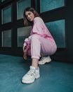 Full body of cheerful ethnic stylish woman in pink outfit and white sneakers hunkering down near black wall with frames and looking at camera in soft daylight