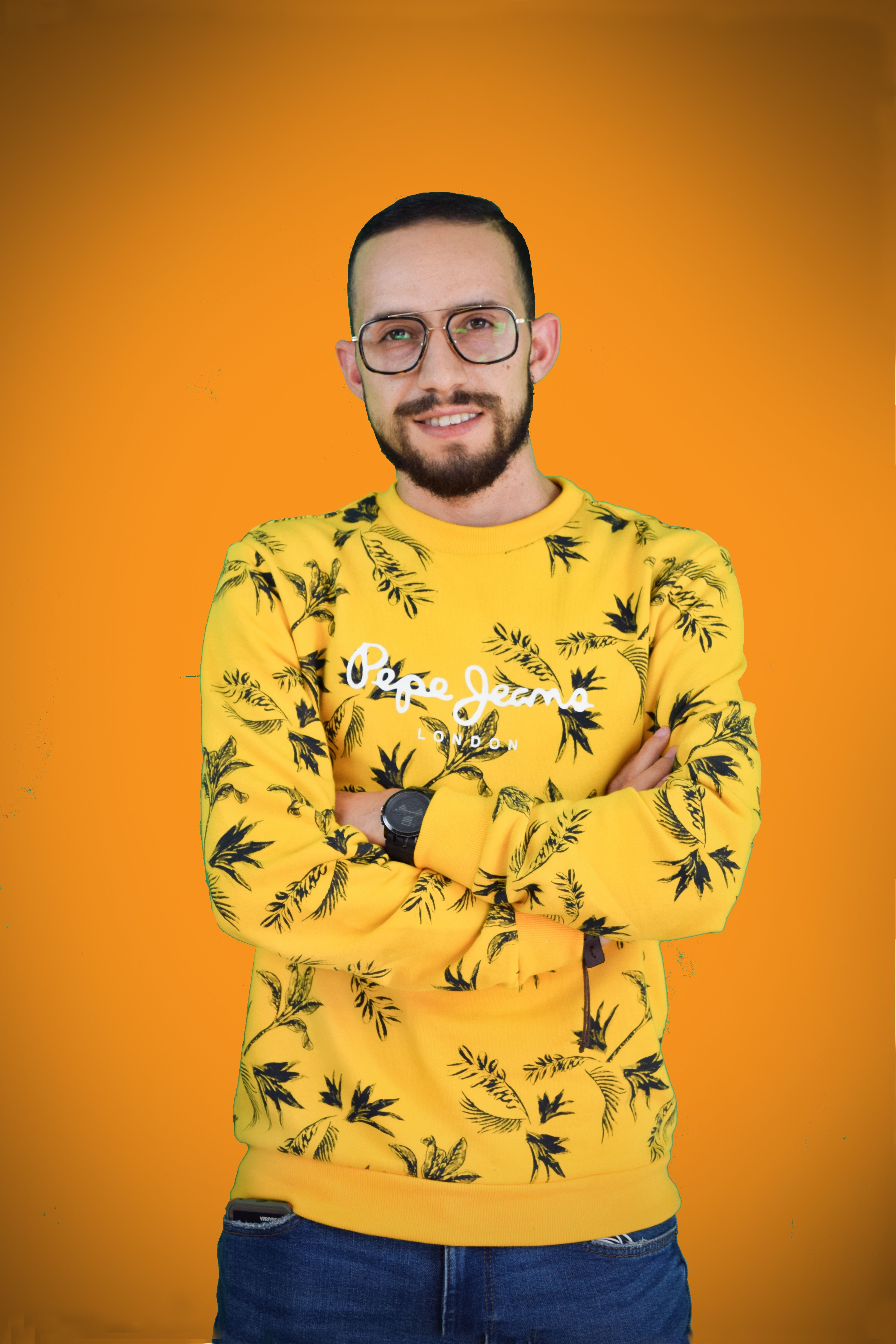 Portrait of a smiling bearded man with glasses, arms crossed in a yellow sweater against an orange background.