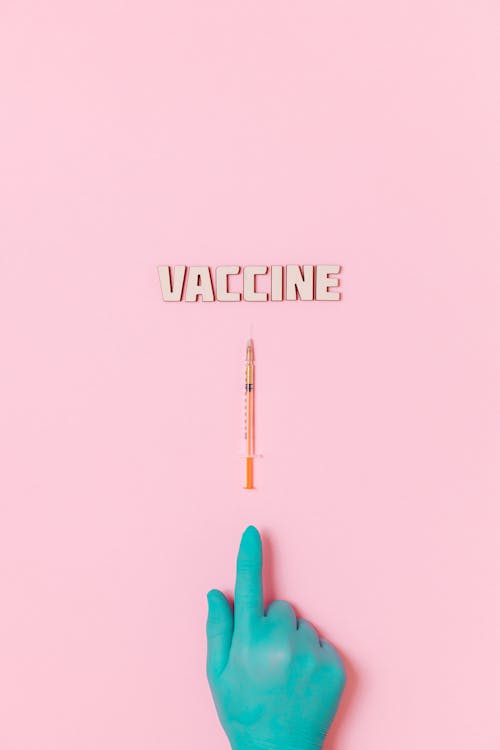 Free A Person's Hand With Latex Glove Pointing His Index Finger to the Syringe and Vaccine Text on Pink Background Stock Photo
