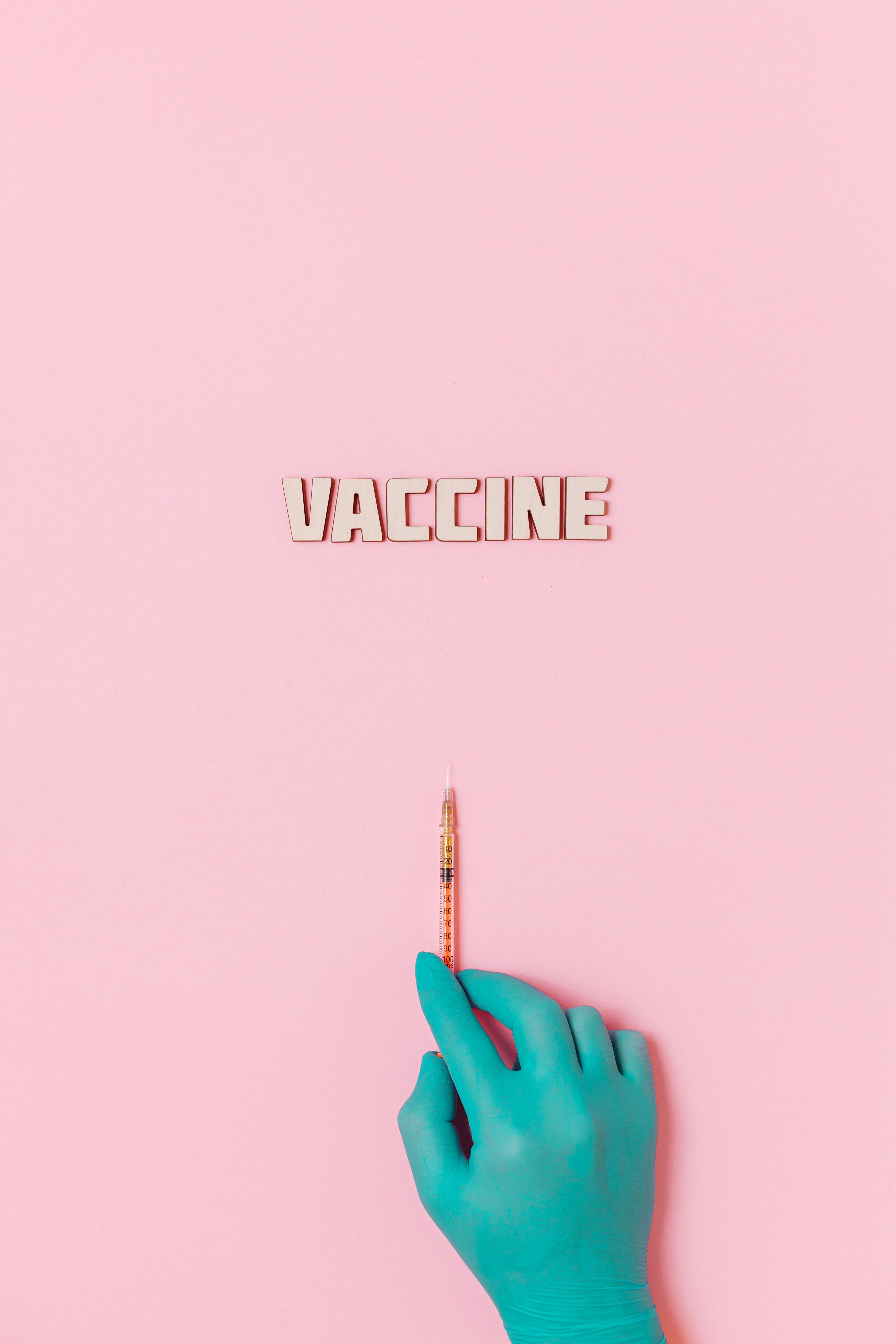 vaccine text and a person wearing latex glove while holding a syringe on pink background