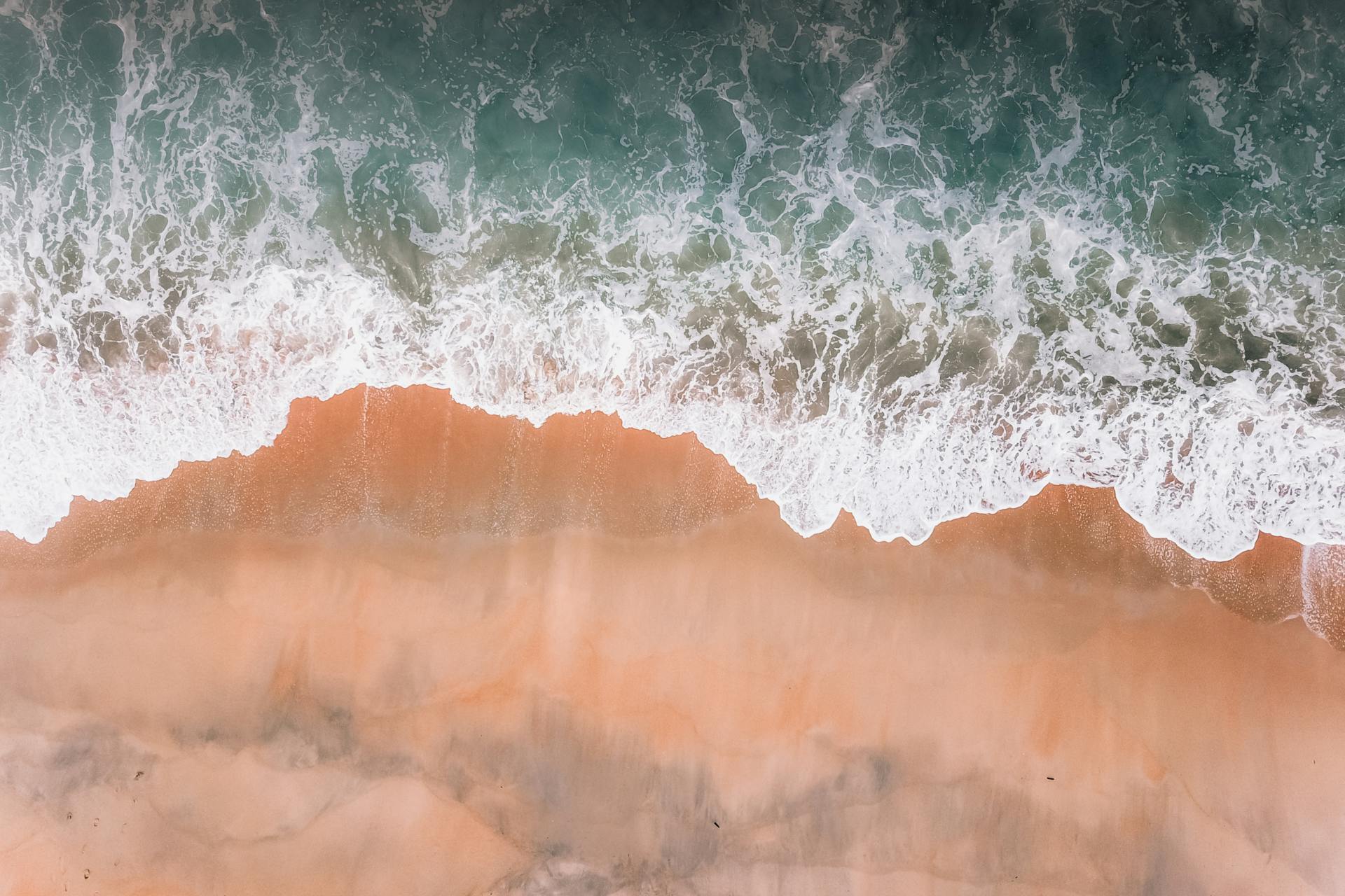 Drone view of waves with foam of deep blue ocean running against sandy coast in soft daylight