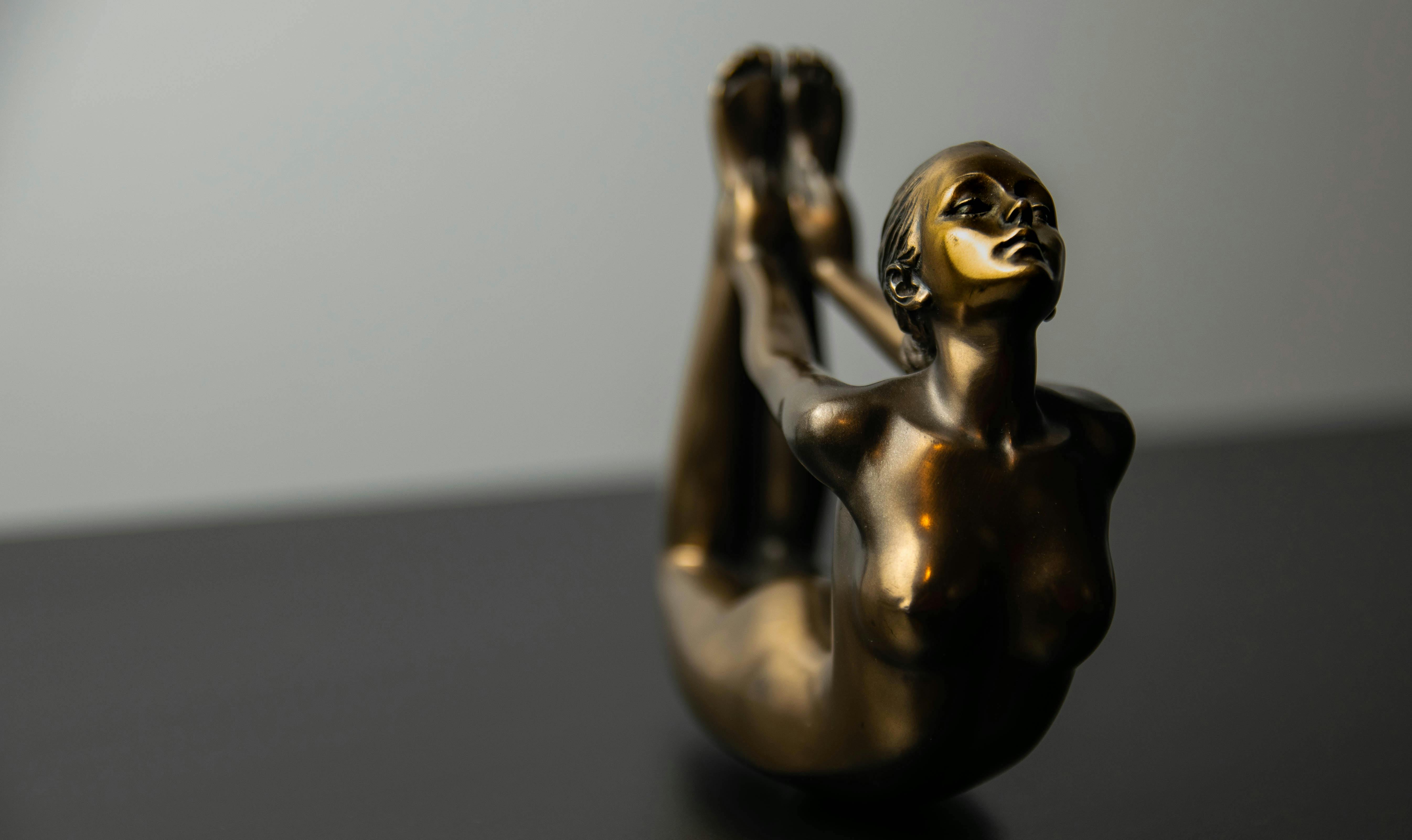 Bronze sculpture of a stretching female figure on a grey background. Modern art piece in a studio setting.