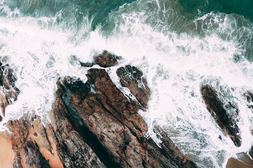 Aerial picturesque view of foamy crystal blue waves of sea splashing against rocky formations in soft daylight