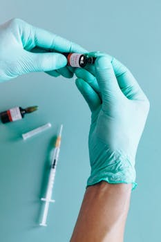 Close-up of hands in gloves handling a vaccine vial and syringe on a teal background.