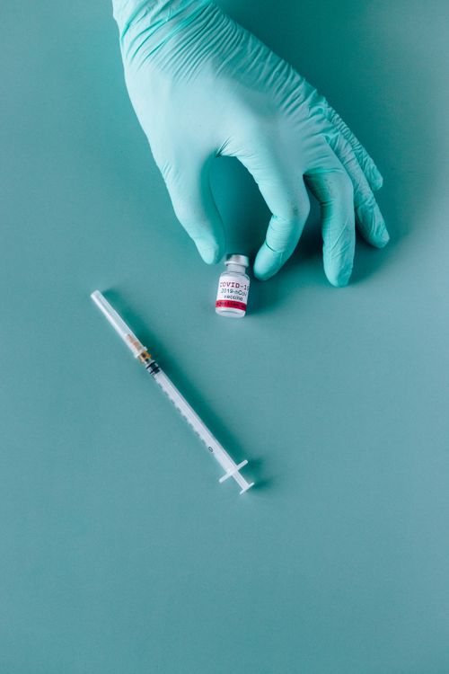 Free A gloved hand holding a COVID-19 vaccine vial next to a syringe on a teal background. Stock Photo